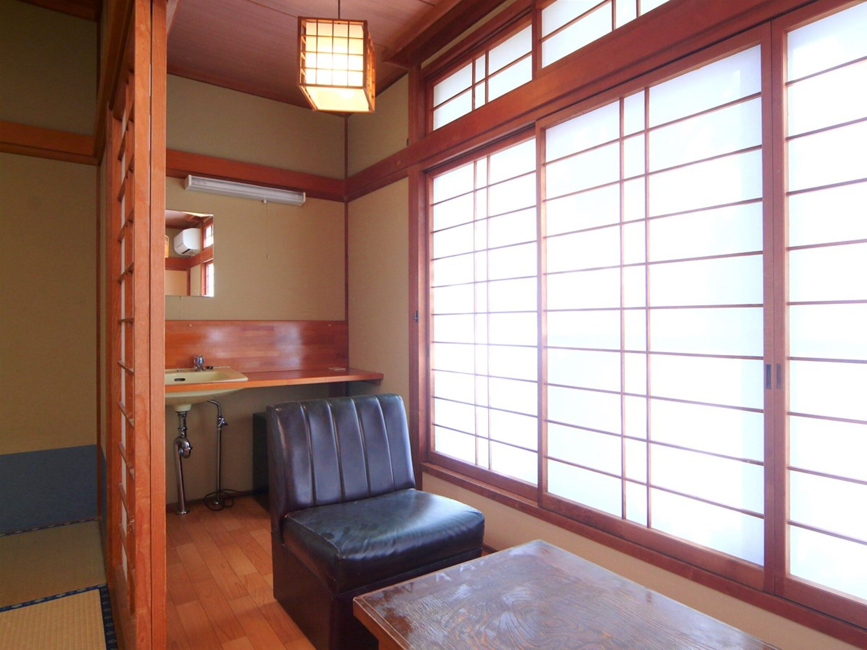 Una habitación japonesa tradicional con suelo de tatami, una mesa de madera baja, un banco acolchado, un lavabo pequeño y una ventana con marcos de madera.