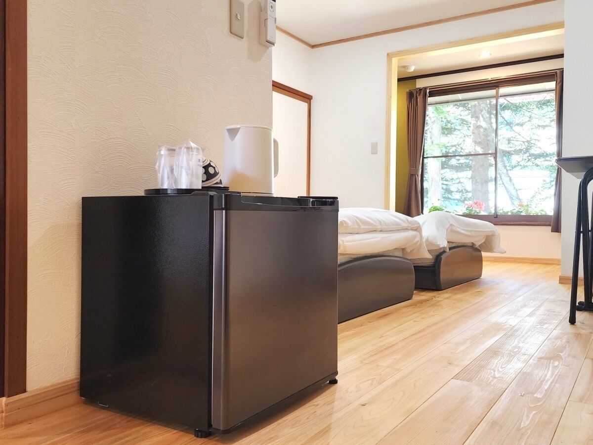 A compact room with a bed, a small black refrigerator, a window with curtains, and a wooden floor.