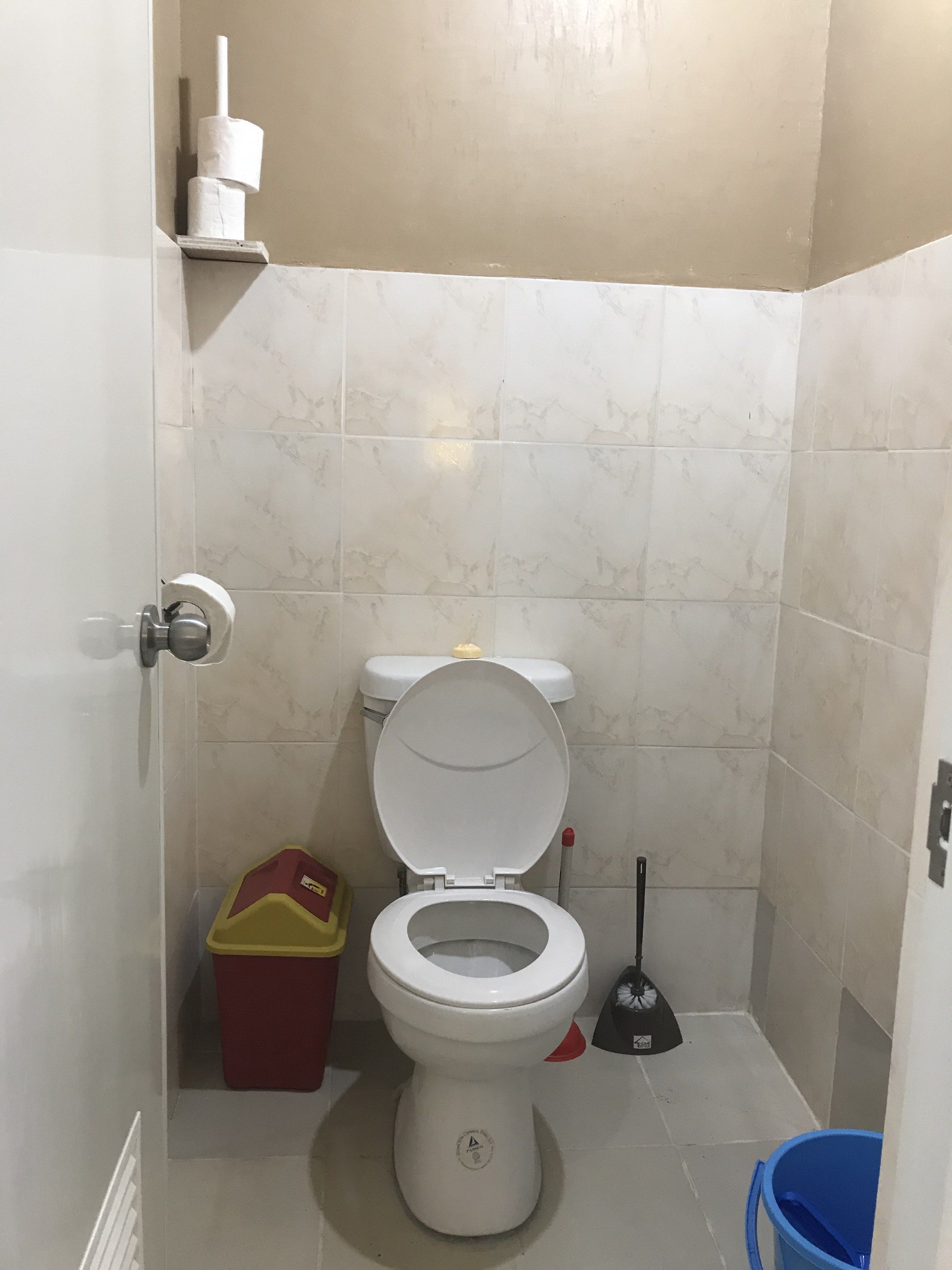 A small bathroom with a toilet, a red trash bin, a blue bucket, and a broom.