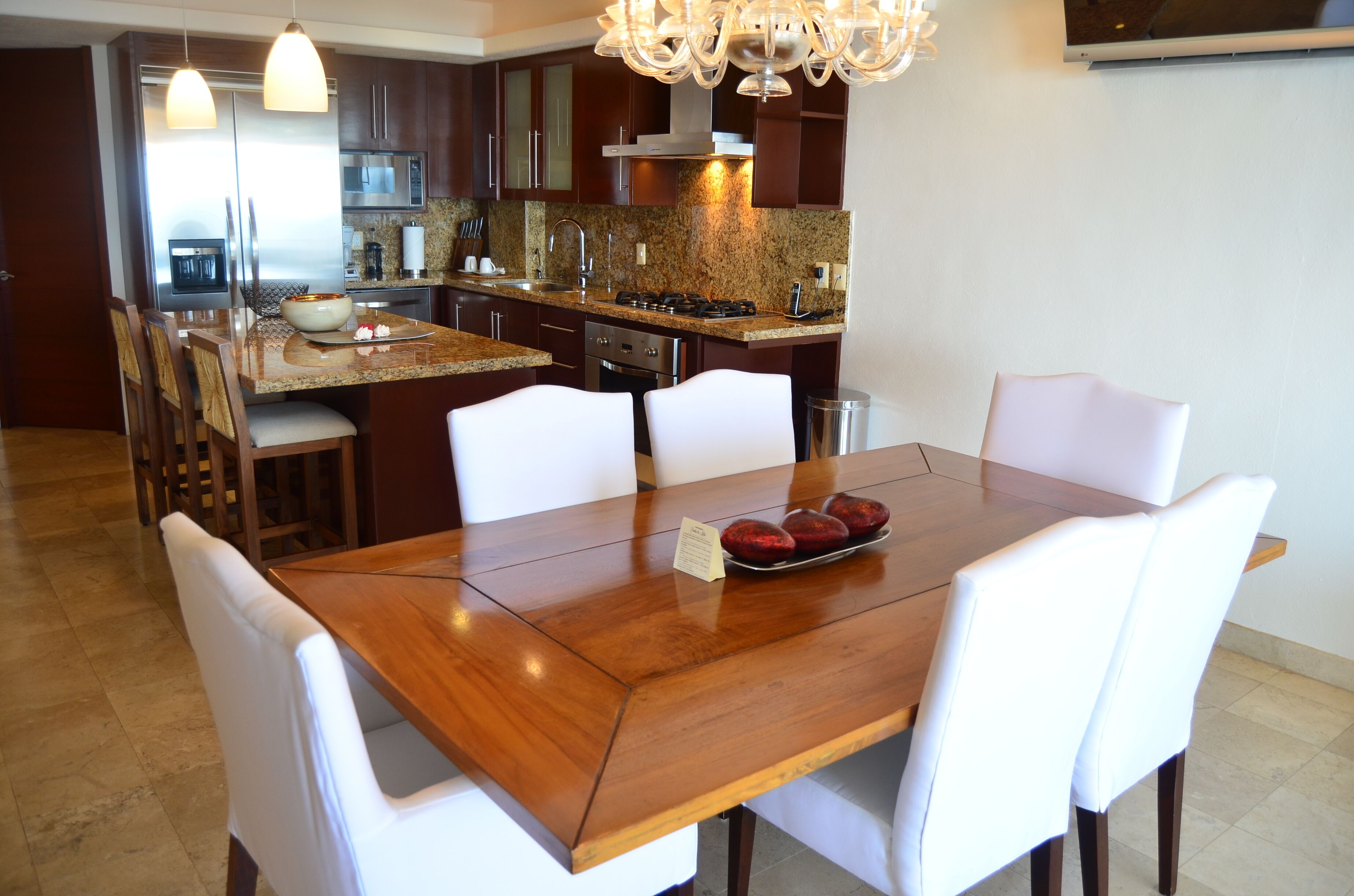 Cocina moderna con armarios de madera oscura, electrodomésticos de acero inoxidable y encimera de granito. Hay una mesa de comedor con sillas blancas y un recipiente pequeño con bayas rojas encima.