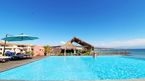 Una piscina azul cristalina con sillas de descanso y sombrillas, una estructura con techo de paja y vistas al mar.