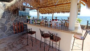 Un área de bar con pared de piedra, techo de paja y vista al océano.