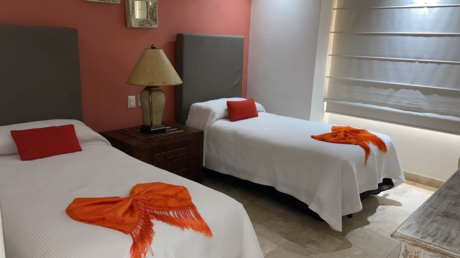 A hotel room with two beds, each with a white bedspread and red pillows. There is a nightstand with a lamp between the beds.