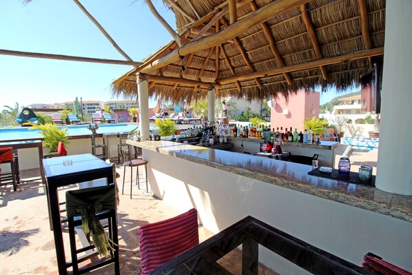Zona de bar con piscina al fondo, techo de paja y asientos al aire libre.