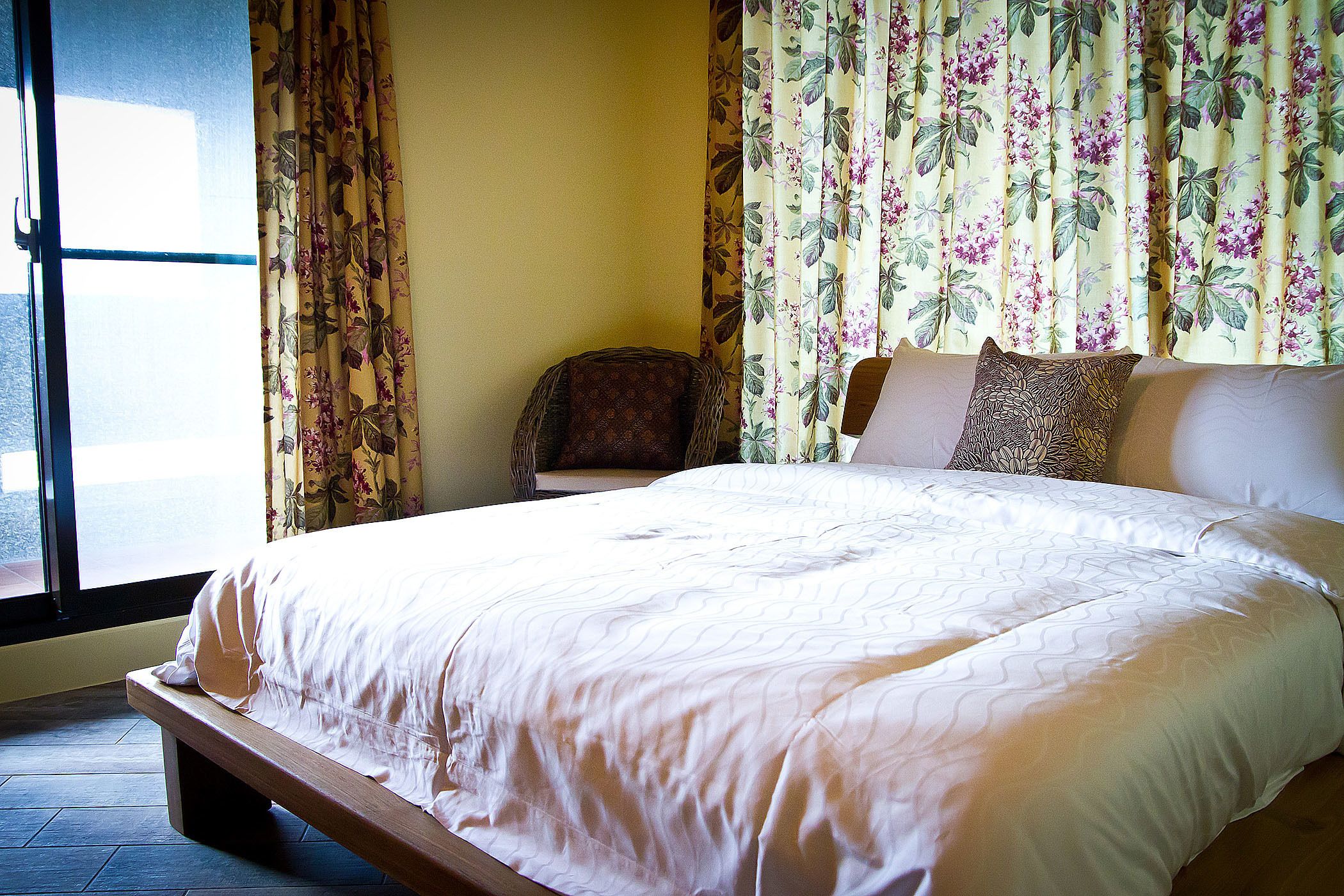 A neatly made bed with a wooden frame, a chair with a patterned cushion, and floral curtains.
