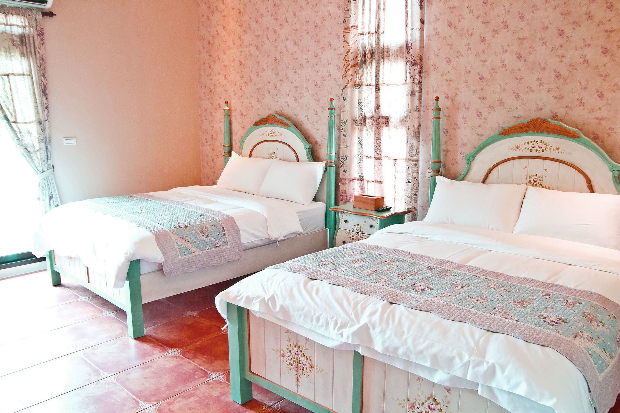 Dos camas individuales con cabeceros iguales, una mesita de noche con lámpara y una ventana con cortinas.
