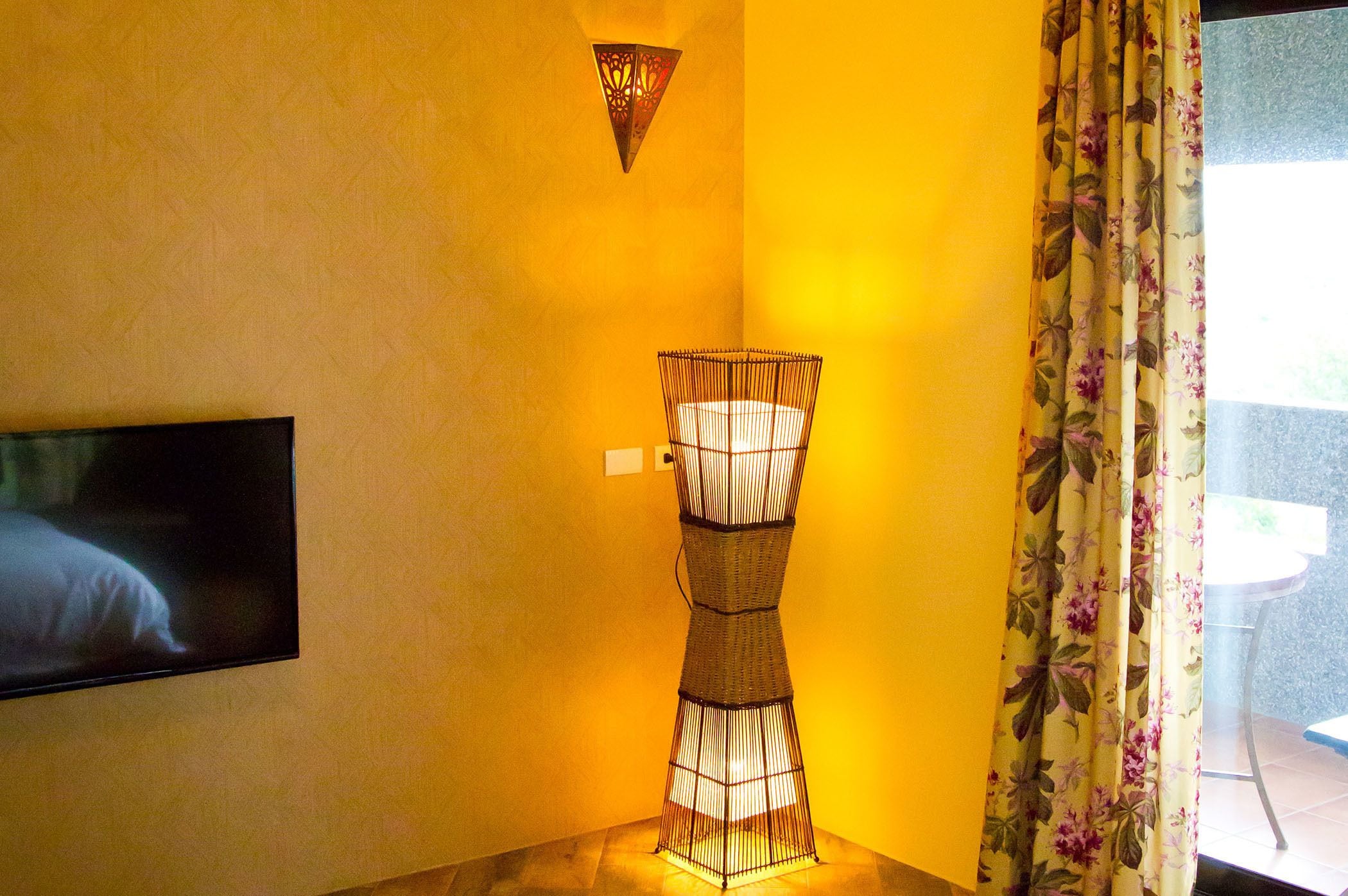 Une pièce avec un téléviseur fixé au mur, une lampe en osier et une fenêtre ornée de rideaux à motifs floraux.