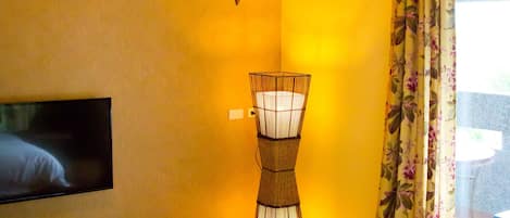 Une pièce avec un téléviseur fixé au mur, une lampe en osier et une fenêtre ornée de rideaux à motifs floraux.