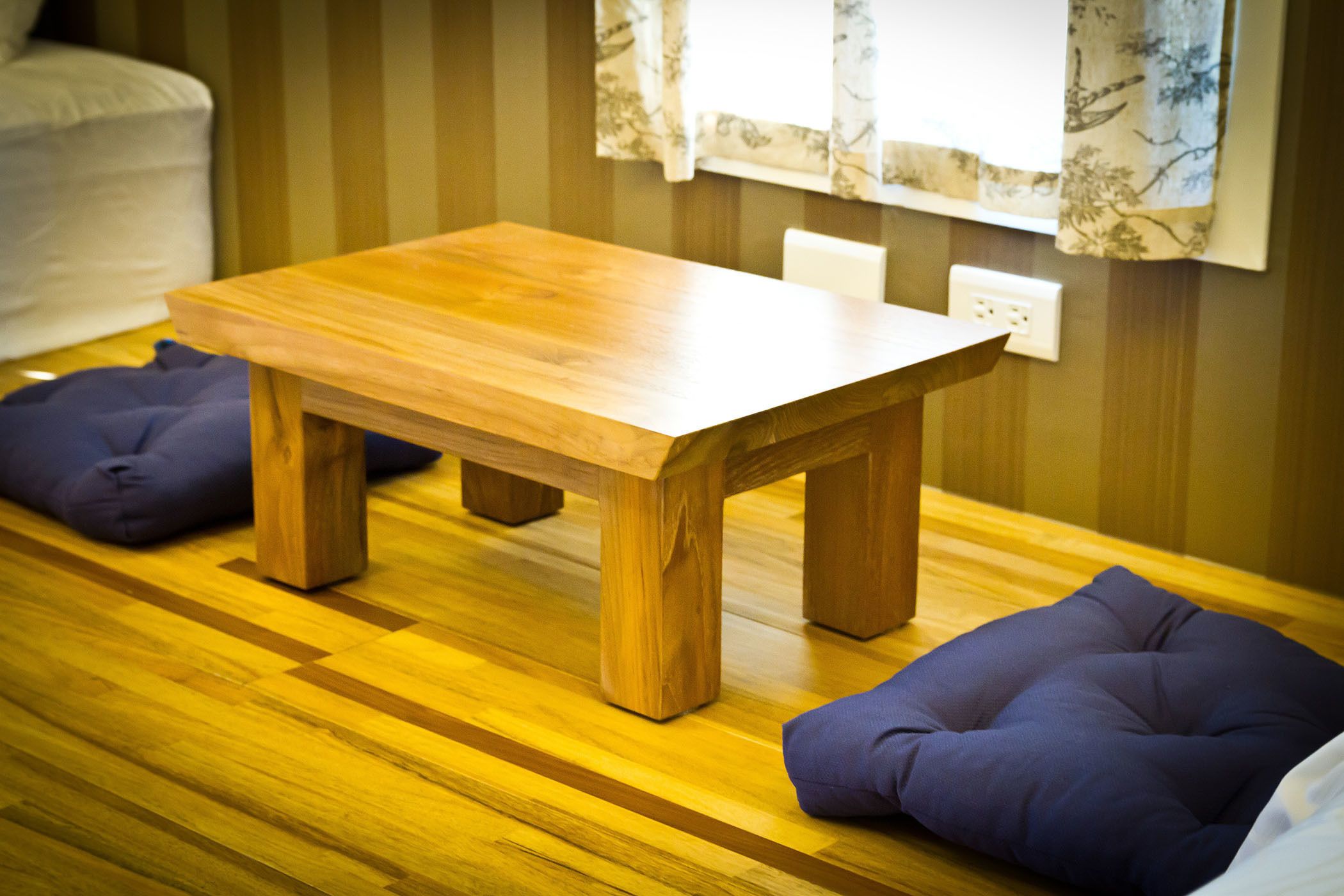 Une table en bois, avec un coussin de chaque côté, posée sur un plancher en bois, le tout devant un mur à rayures.