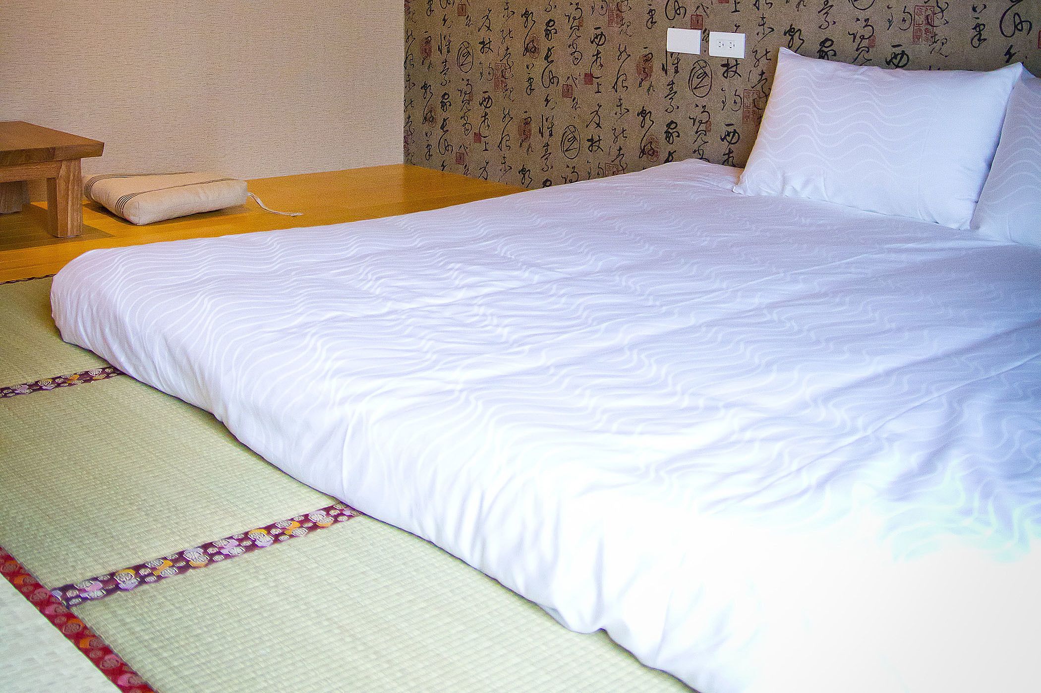 A neatly made bed with white bedding, a wooden nightstand, and a tatami mat.