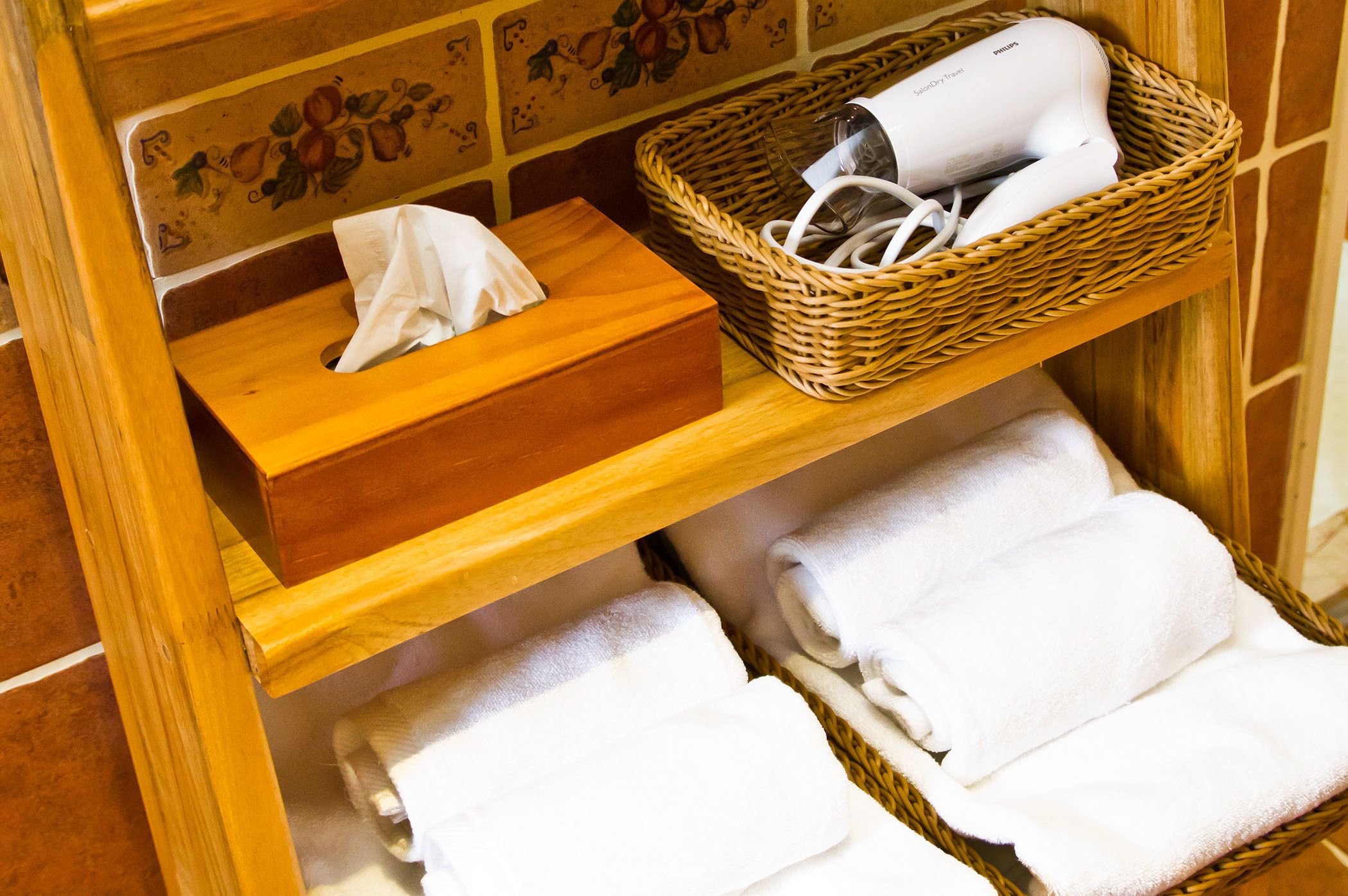 A wooden shelf with a tissue box, a basket containing a hairdryer, and neatly folded towels.