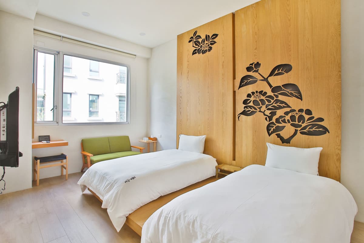 Habitación de hotel con dos camas, un muro decorativo de madera con diseño floral, un sofá verde, una mesita y una ventana con vistas a edificios.