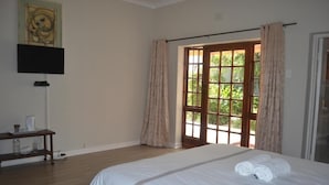 A hotel room with a large bed, a flat-screen TV mounted on the wall, a small table with a lamp, and a window with curtains. - Cycad Lodge (East London)