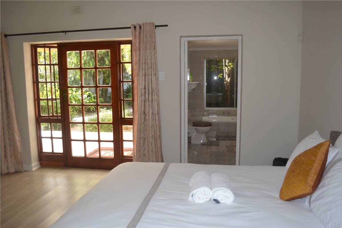 A hotel room with a large bed, a bathroom with a bathtub, and a sliding glass door leading to a garden.