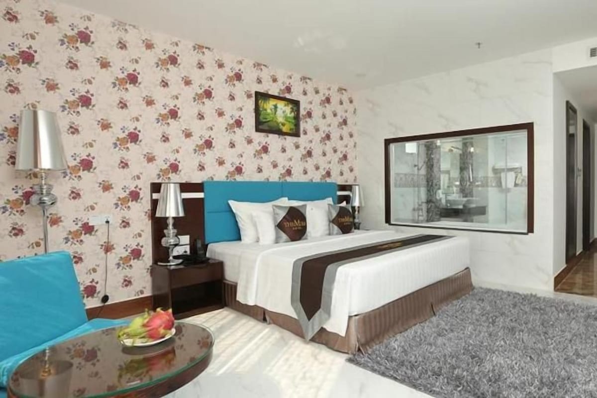 A hotel room with a bed, a nightstand, a lamp, a small table with a fruit bowl, a grey rug, and a large window.