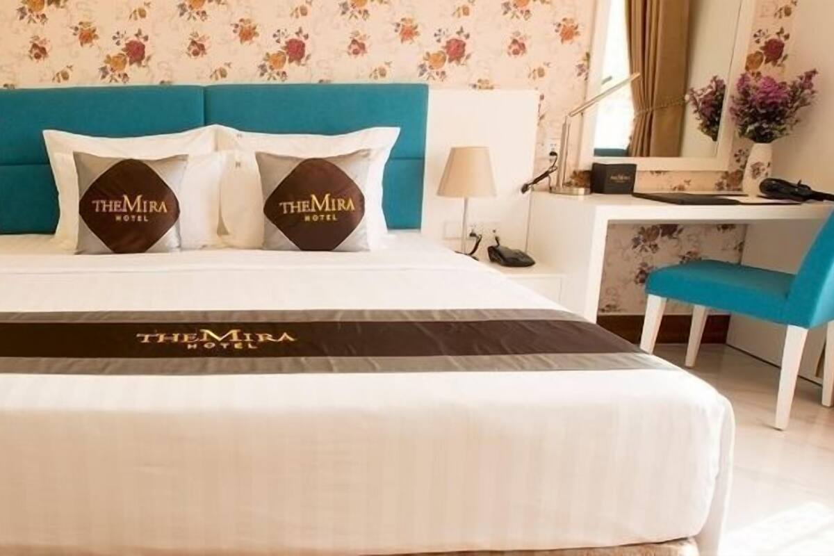 Une chambre d’hôtel avec un grand lit, un bureau et une chaise. Le lit est doté d’une tête de lit et de deux oreillers ornés du logo de l’hôtel.
