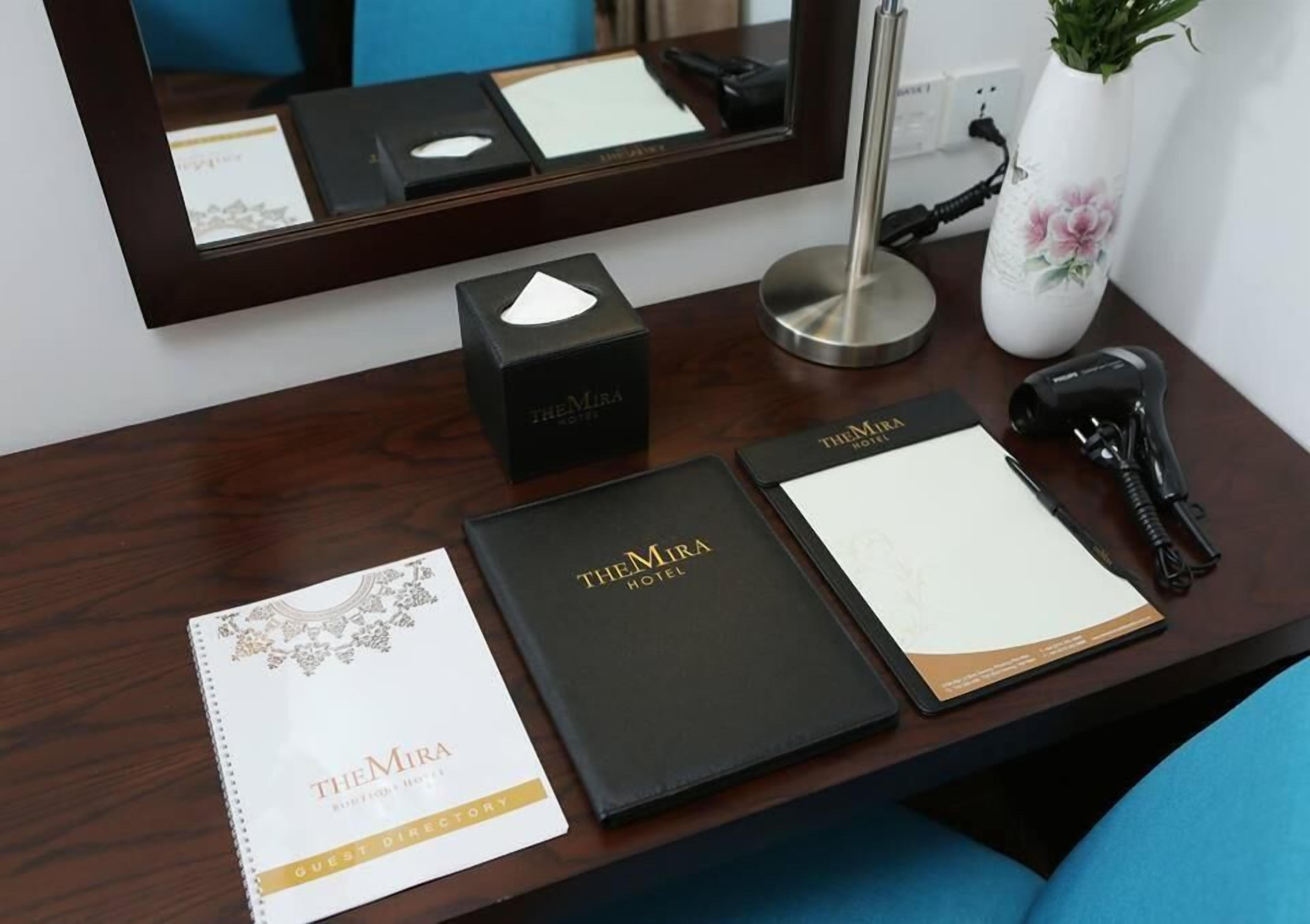 Un mostrador de hotel con un directorio para huéspedes, una carpeta negra con la etiqueta "TheMira Hotel", una caja negra, un jarrón blanco con una flor rosa, una lámpara de escritorio y un teléfono negro.