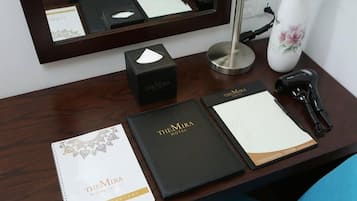 Un mostrador de hotel con un directorio para huéspedes, una carpeta negra con la etiqueta "TheMira Hotel", una caja negra, un jarrón blanco con una flor rosa, una lámpara de escritorio y un teléfono negro.