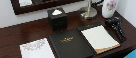 Un mostrador de hotel con un directorio para huéspedes, una carpeta negra con la etiqueta "TheMira Hotel", una caja negra, un jarrón blanco con una flor rosa, una lámpara de escritorio y un teléfono negro.