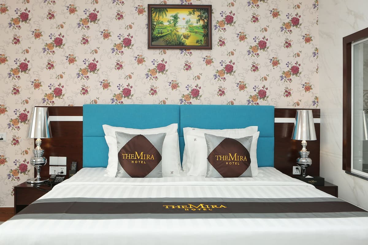Une chambre d’hôtel avec un papier peint à motifs floraux, un tableau représentant un paysage encadré, une tête de lit bleue et des oreillers ornés du nom de l’hôtel.