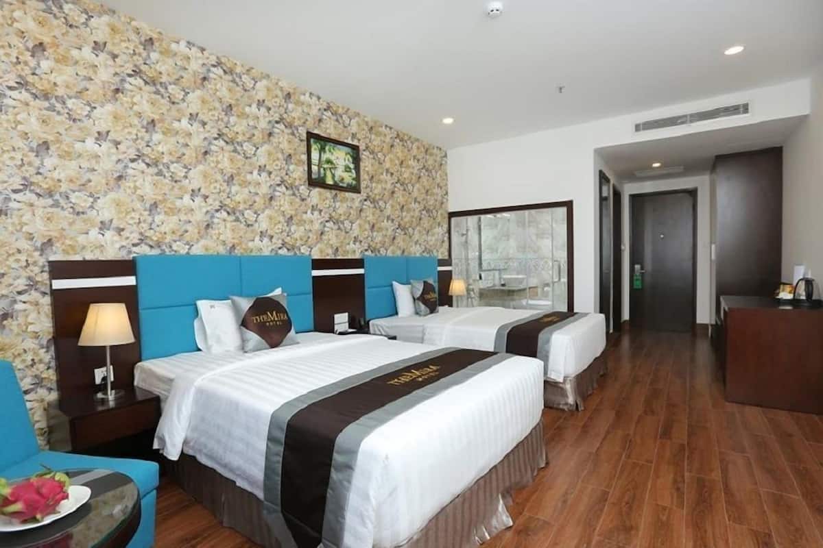 Une chambre d’hôtel avec deux lits, un mur décoré de motifs floraux, un bureau et une salle de bain visible à travers une porte en verre.