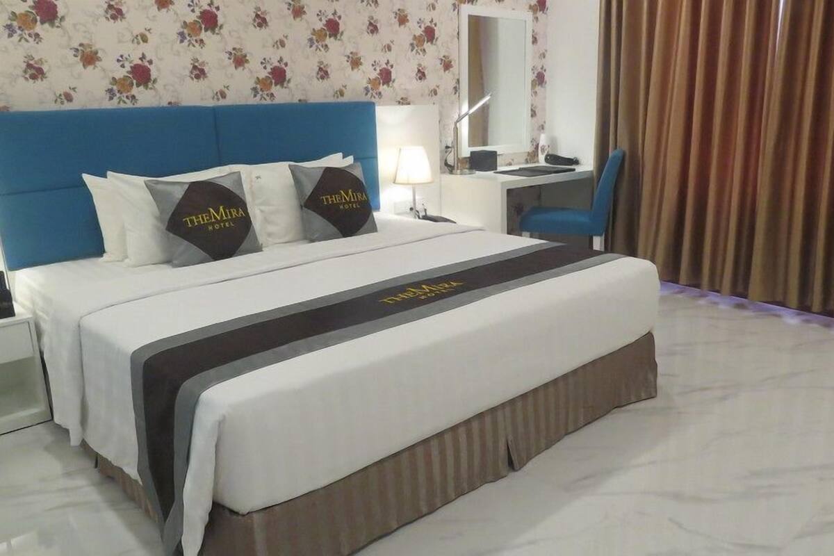 Une chambre d’hôtel avec un lit, un bureau, une chaise, une lampe et une fenêtre avec des rideaux.