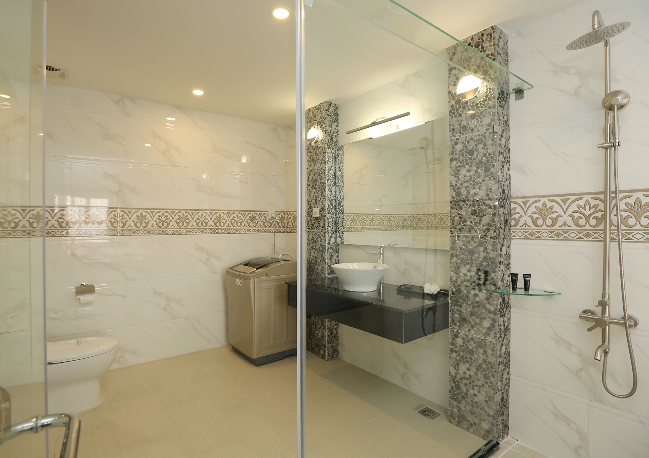 A modern bathroom with a glass-enclosed shower, a marble-tiled wall, a sink with a glass shelf, and a toilet.