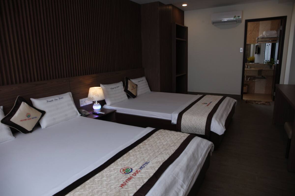 Une chambre d’hôtel avec deux lits, une table de chevet avec une lampe, une tête de lit en bois, un miroir et une petite kitchenette.