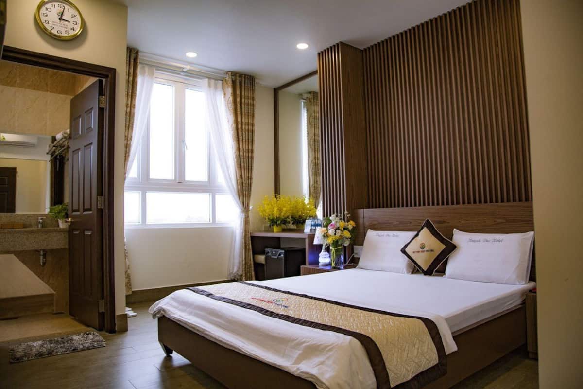 Une chambre d’hôtel avec un grand lit, une tête de lit en bois, une table de chevet ornée d’un vase de fleurs et une fenêtre avec des rideaux.