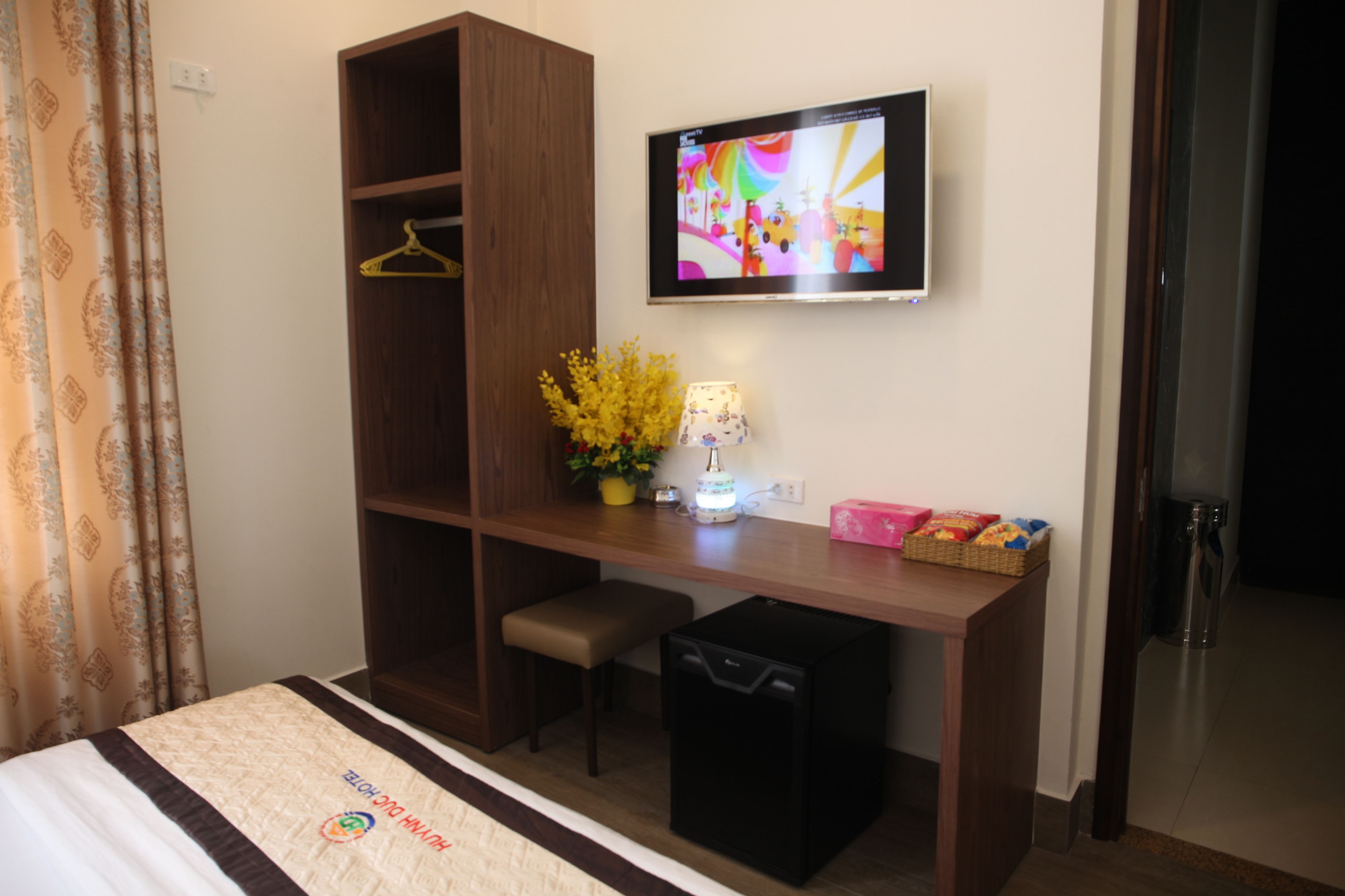 A hotel room with a bed, a desk, a TV, a small bench, a trash bin, and a wall-mounted TV.