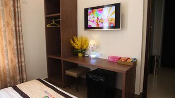 A hotel room with a bed, a desk, a TV, a small bench, a trash bin, and a wall-mounted TV.