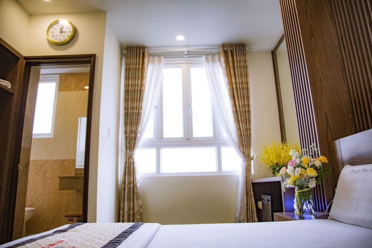 Une chambre d’hôtel avec un lit, une horloge au mur, une fenêtre avec des rideaux et un vase rempli de fleurs.