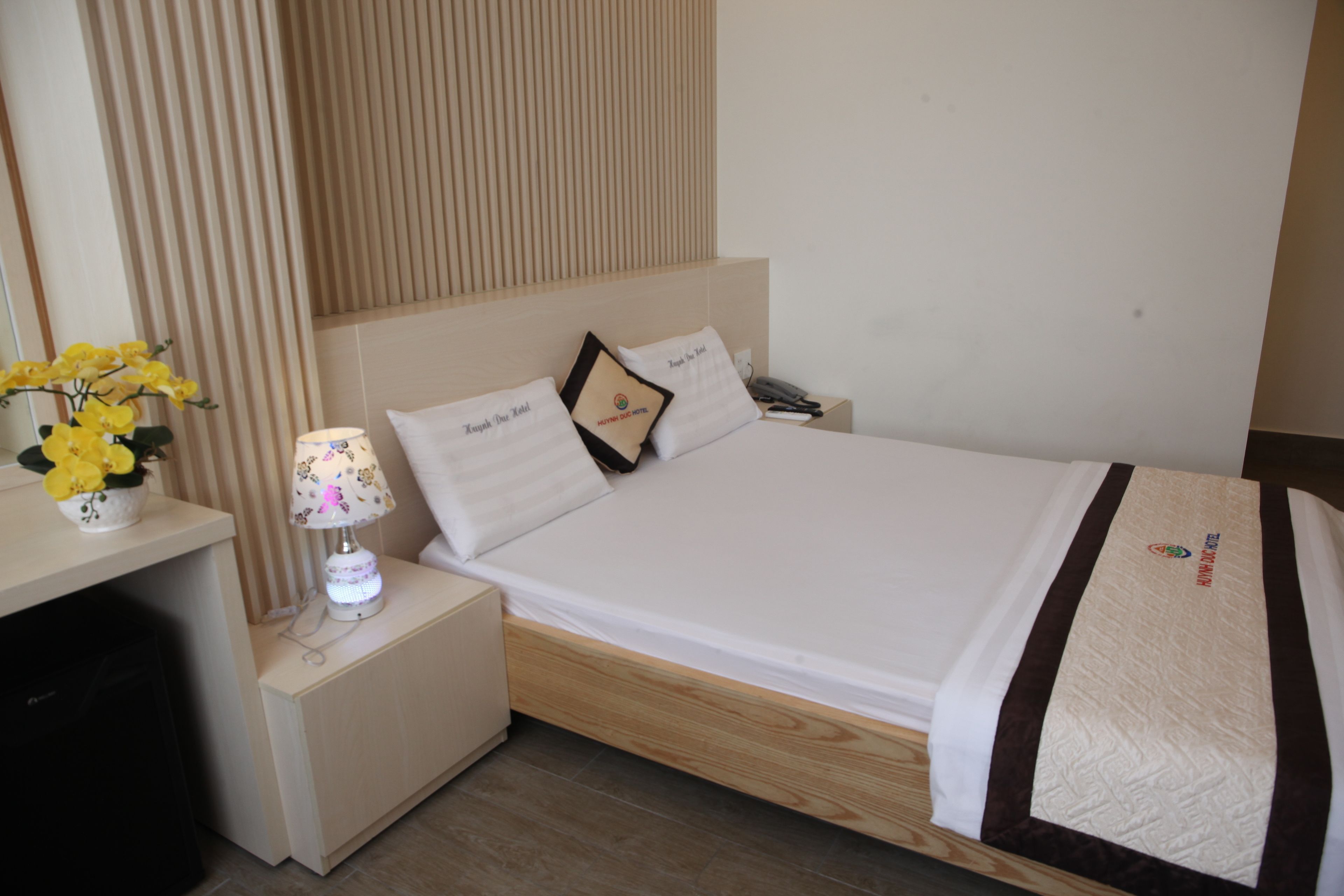 A neatly made bed with a white and brown bedspread, a bedside lamp, and a small vase with yellow flowers on a bedside table.
