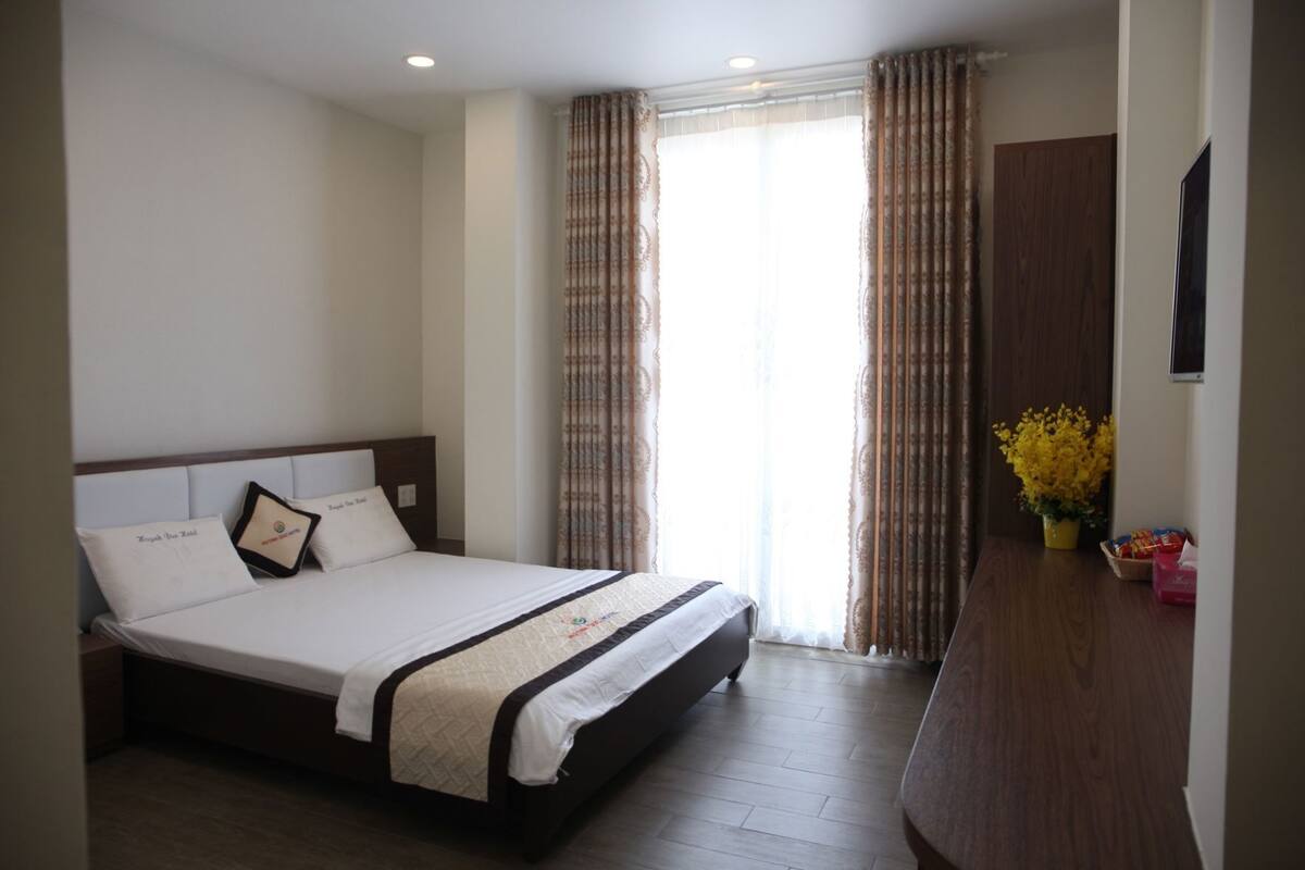 Une chambre d’hôtel avec un lit, des rideaux, un bureau en bois et une plante en pot.