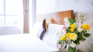 Una cama bien hecha con sábanas blancas y un cabecero de madera, un florero con flores amarillas y blancas sobre la mesita de noche, y una ventana con cortinas.