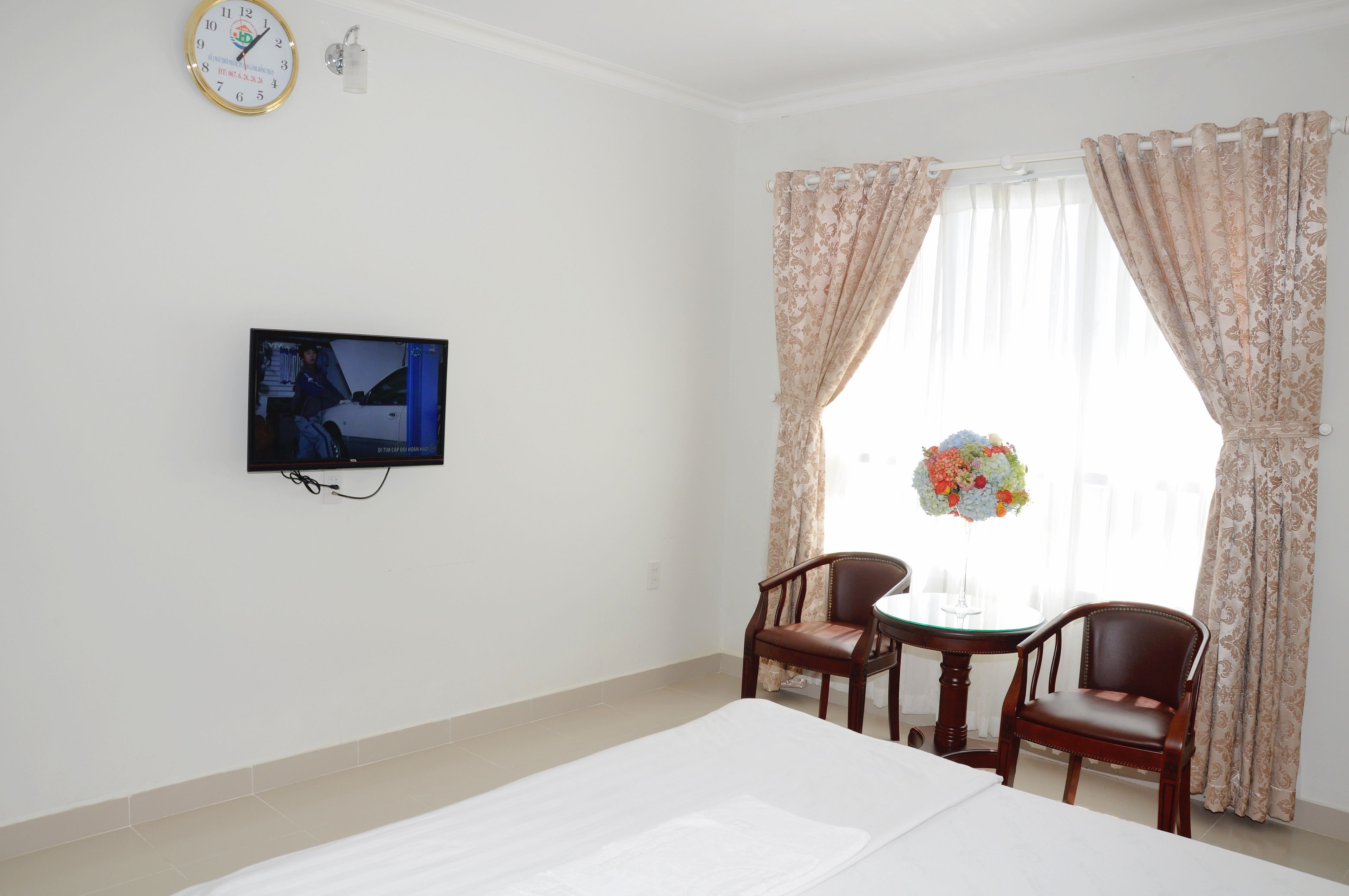 A hotel room with a bed, a TV mounted on the wall, a dining table with two chairs, and a window with curtains.