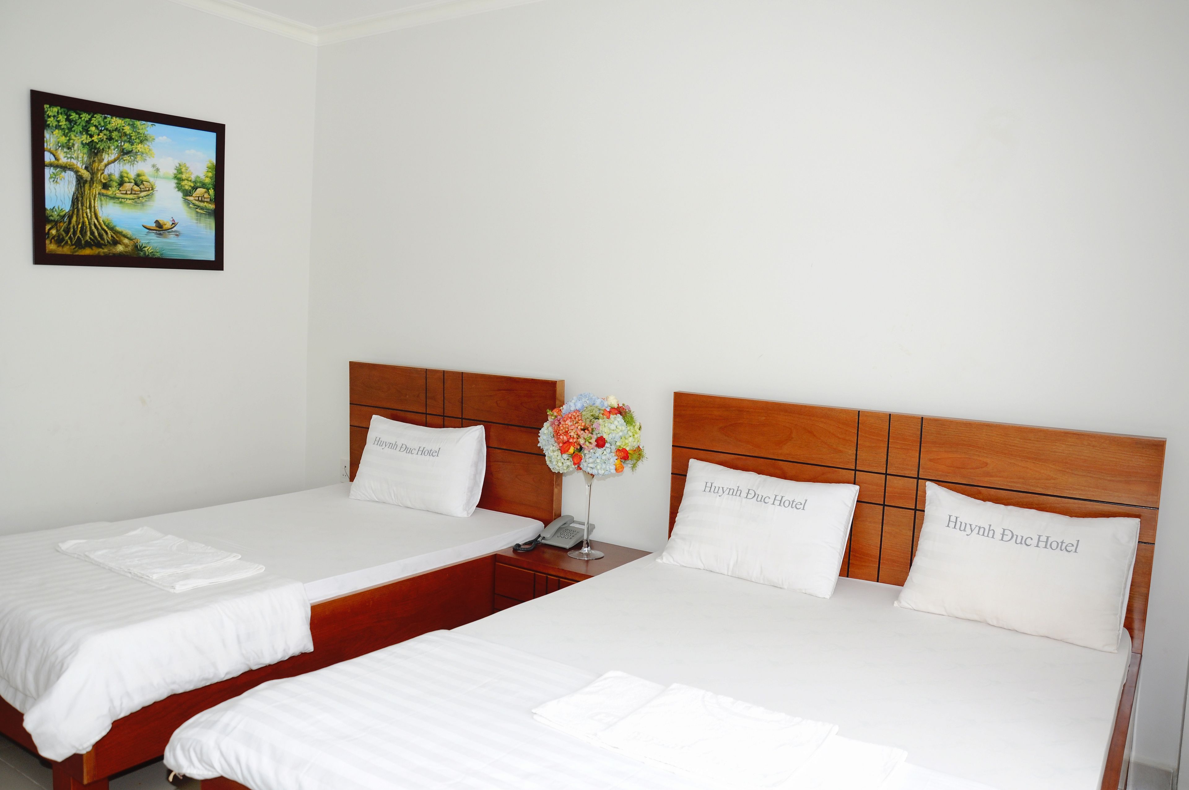 A hotel room with two beds, each with a wooden headboard and white bedding. There is a framed picture on the wall and a vase with flowers on the nightstand.