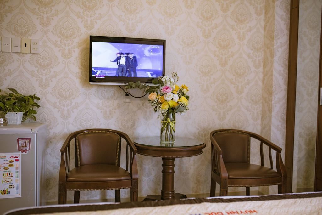Une pièce comprenant deux chaises en bois, une petite table ronde ornée d’un vase de fleurs, un téléviseur fixé au mur et un réfrigérateur sur lequel est affiché un menu.