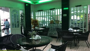A restaurant with glass tables and black wicker chairs, green ceiling lights, and a view through large windows.