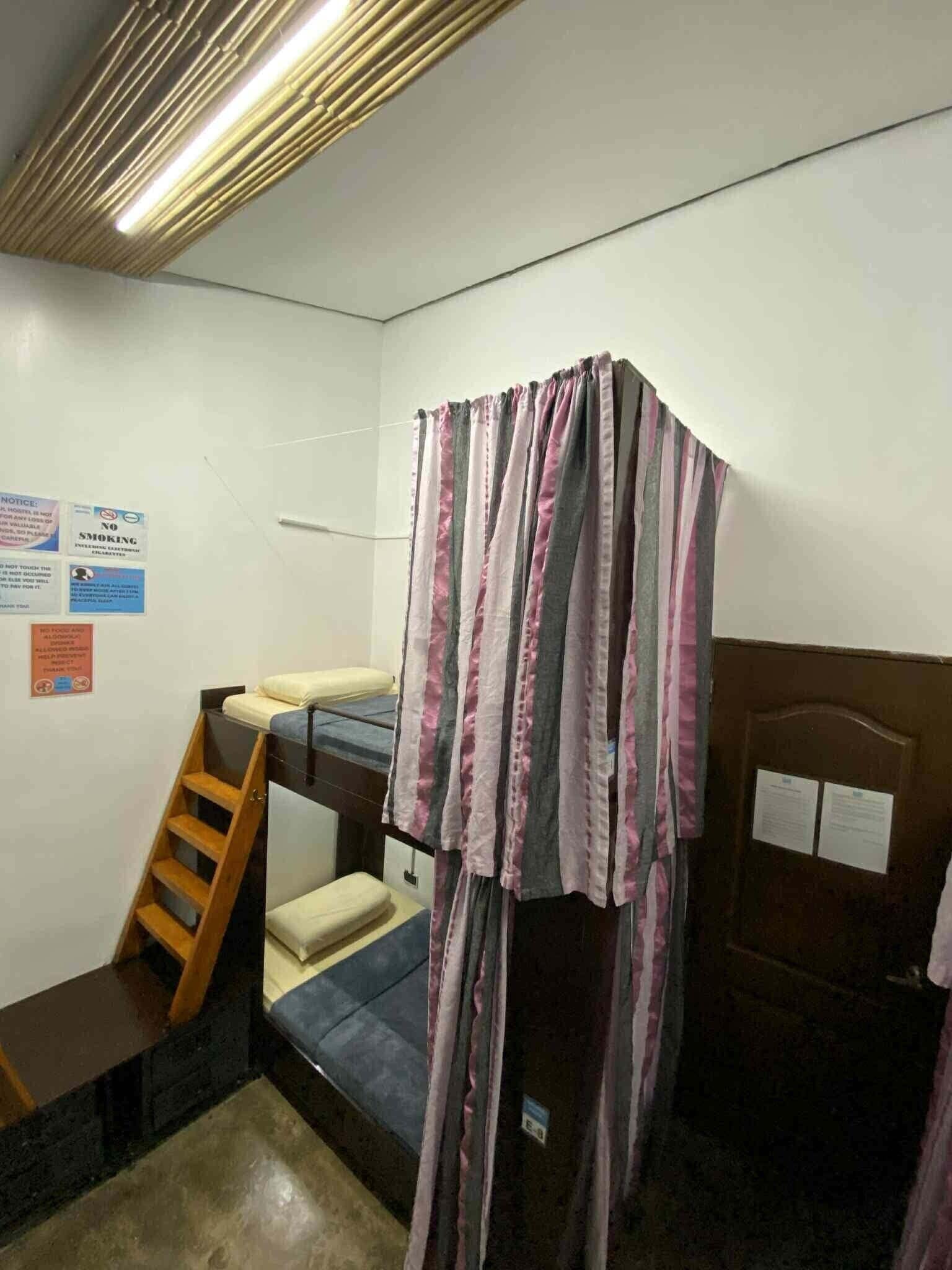Une petite pièce, de taille modeste, avec un lit superposé, une échelle en bois et un rideau. On peut y voir des panneaux sur le mur, dont un panneau indiquant qu'il est interdit de fumer.