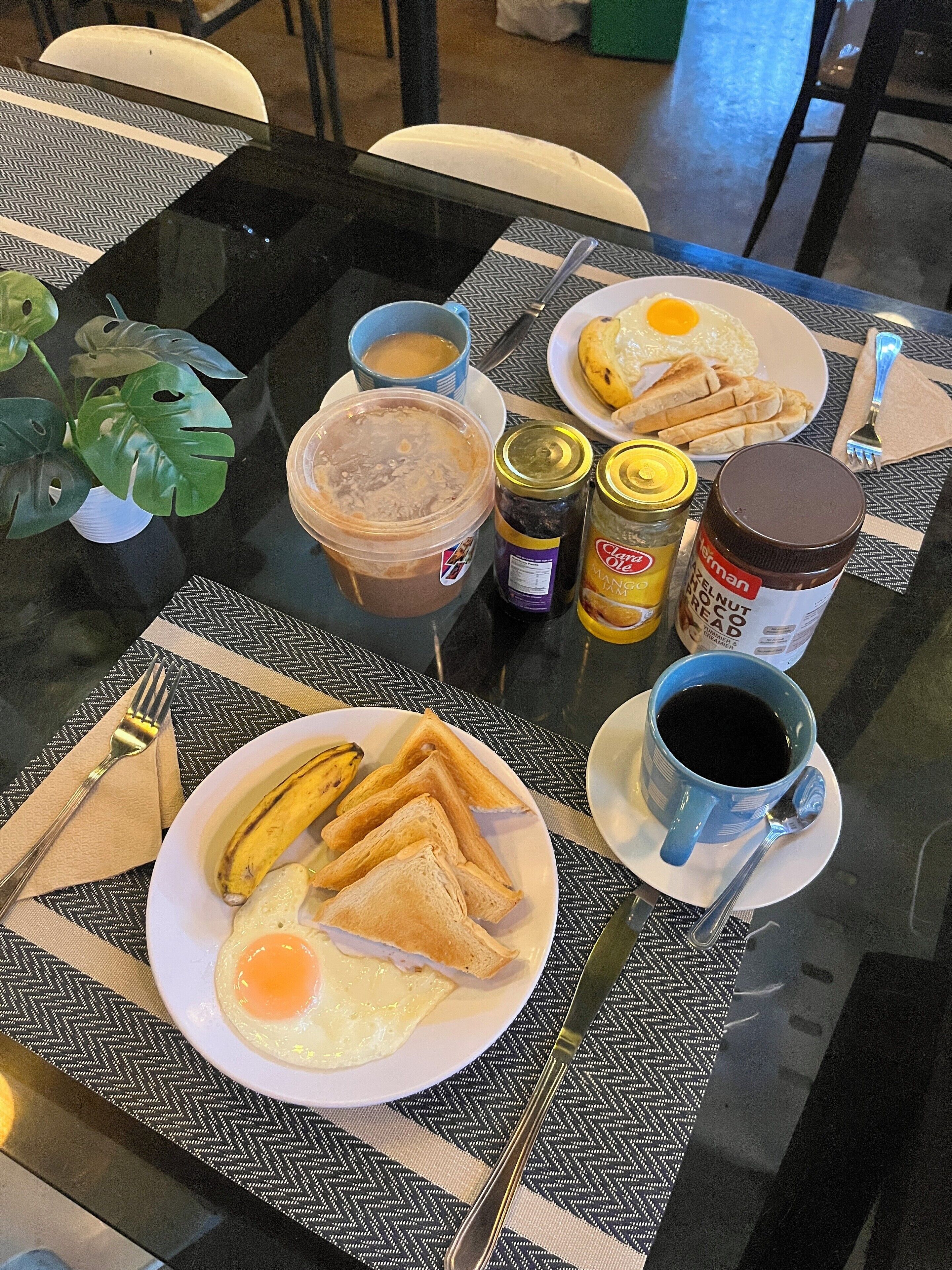 A breakfast table with plates of toast, eggs, and a banana, along with condiments and a coffee cup.