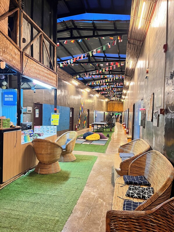 A hostel interior with a high ceiling, colorful flags, wicker furniture, and a green artificial turf area.