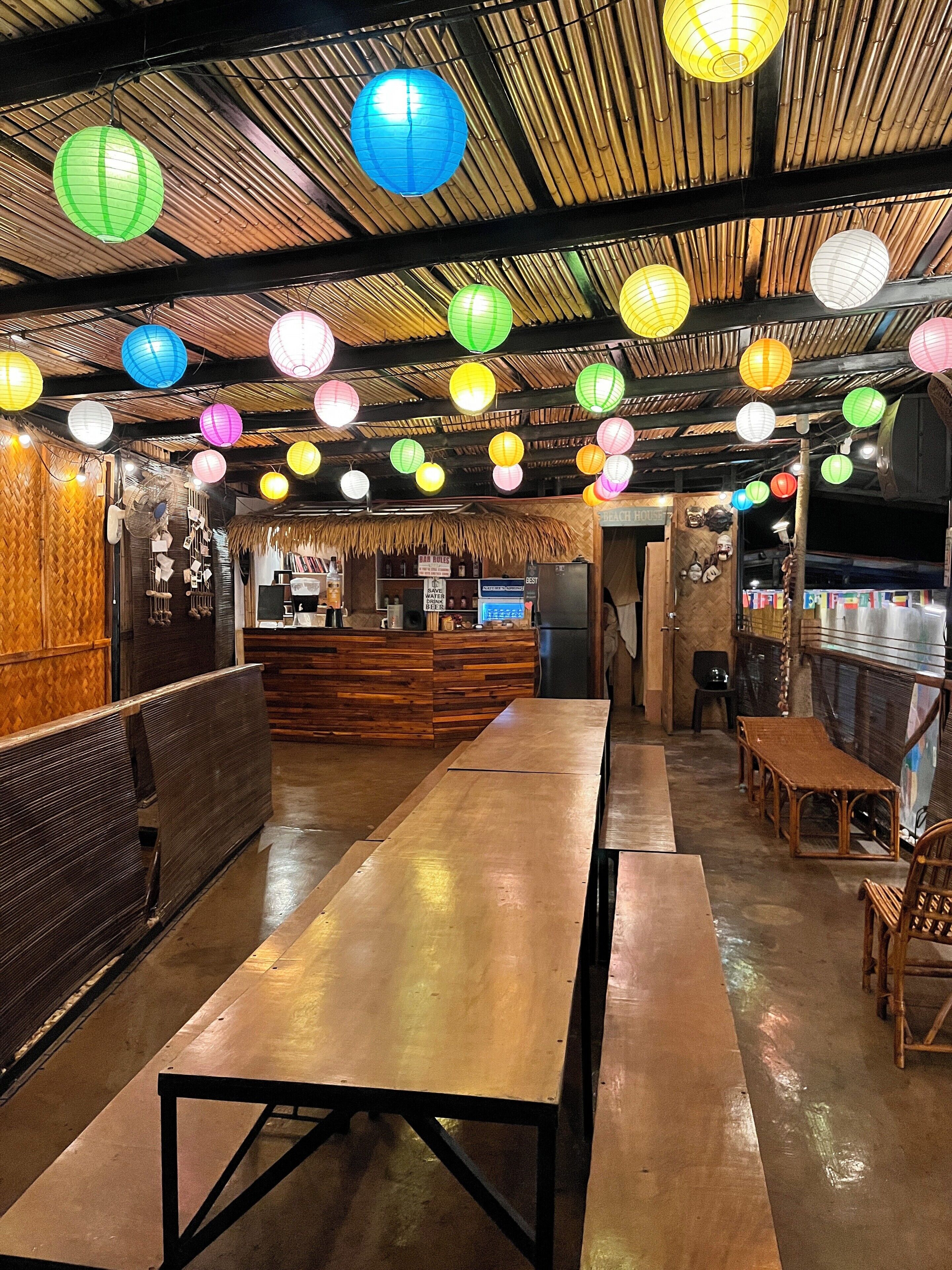 Un restaurante con largas mesas y bancos de madera, una barra con menú y coloridos faroles colgando del techo.