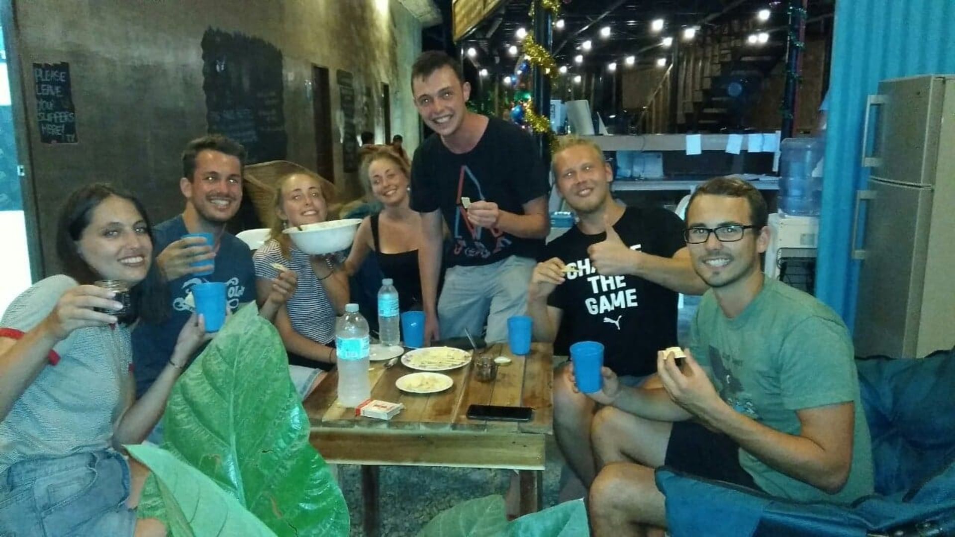 Un grupo de personas sentadas alrededor de una mesa con un bol y botellas de agua, en una habitación con una mesa de madera y sillas azules.