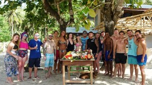 Un groupe de personnes s’est réuni autour d’une table où étaient disposés de la nourriture et des boissons. Elles se trouvaient à l’extérieur, avec des arbres et une structure en bois en arrière-plan.