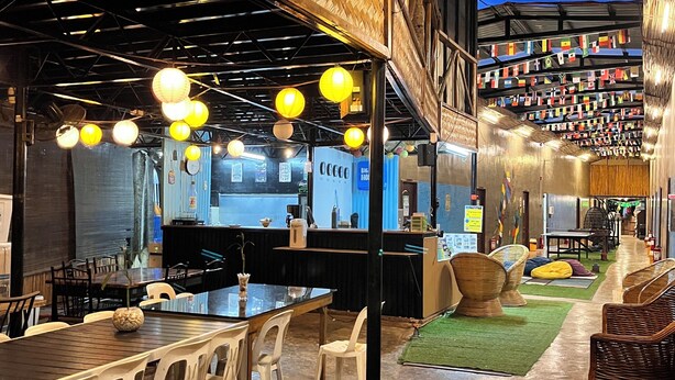 Un restaurant spacieux avec de hauts plafonds, des poutres apparentes et des lanternes suspendues. Les tables et les chaises sont disposées pour accueillir les clients.
