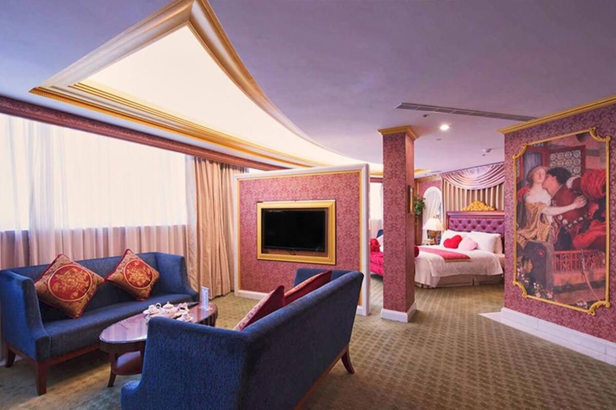 Una habitación de hotel con un conjunto de sofá, un televisor de pantalla plana, una cama con cabecera roja y un cuadro grande en la pared.