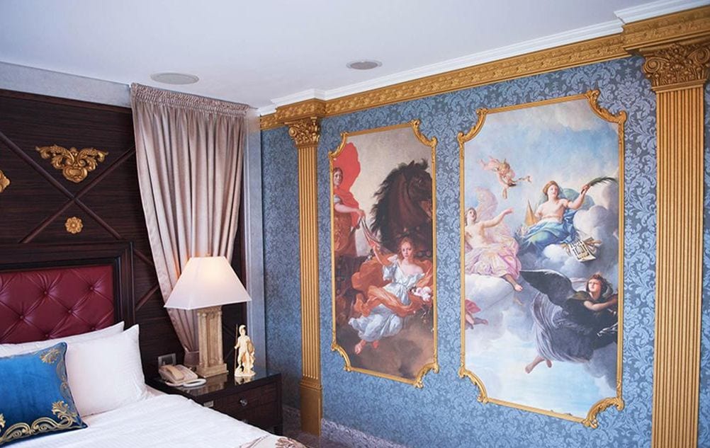 A luxurious bedroom with a large bed, a nightstand, a lamp, and two ornate paintings on the wall.