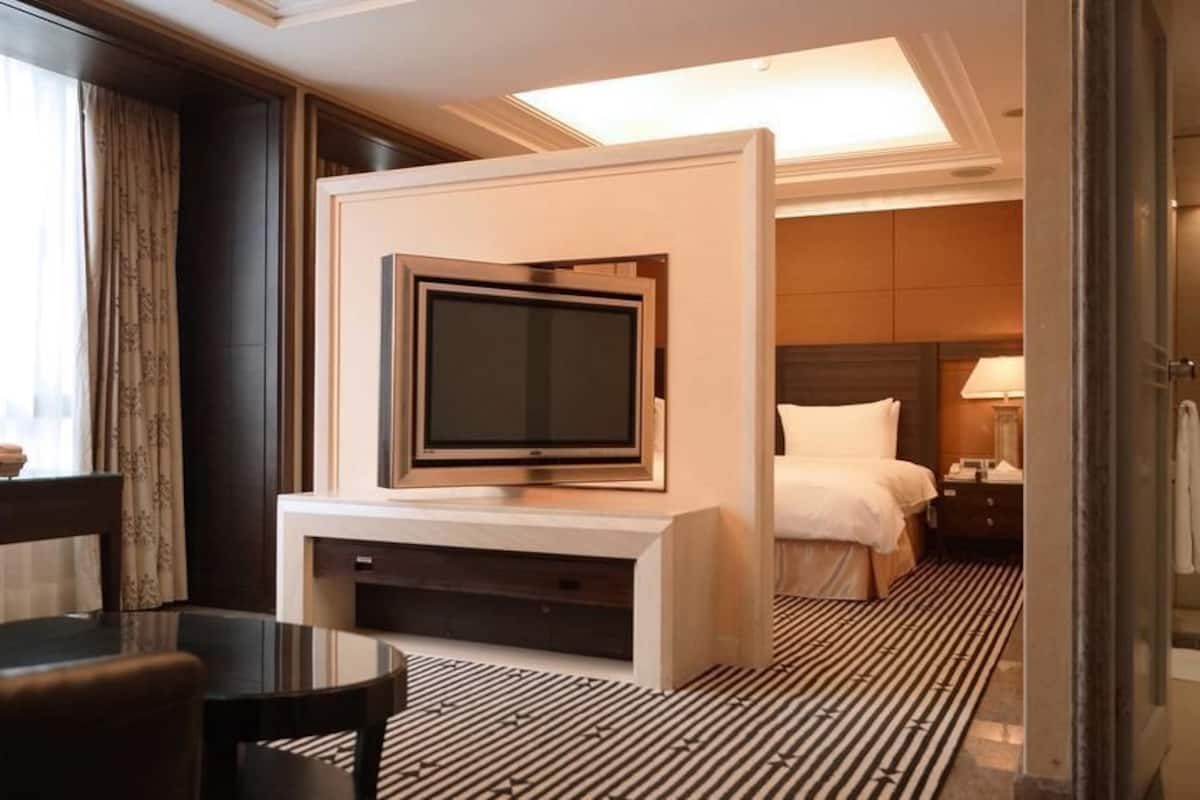 Una habitación de hotel con televisión de pantalla plana en la pared, una cama con ropa blanca, una mesita de noche con lámpara y una alfombra estampada.