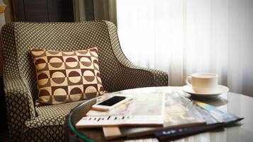 A hotel room with a patterned armchair, a glass coffee table with a magazine, a smartphone, and a cup on a saucer.